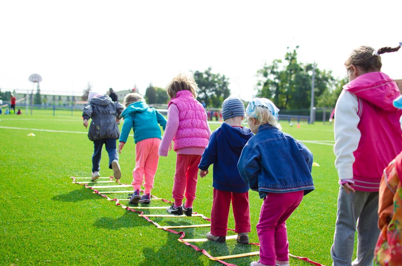 who-we-are Children enjoy an outdoor activity on a grassy field, stepping over a ladder.